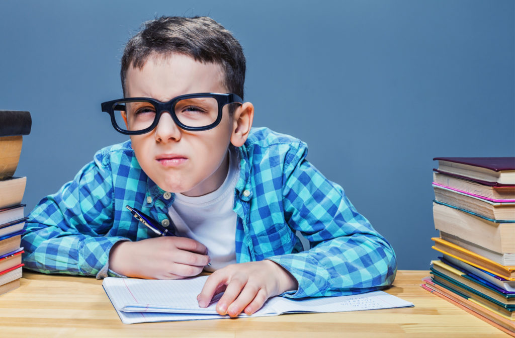 A young child with a stack of books beside him squinting and leaning closer to see more clearly and to be able to take notes.
