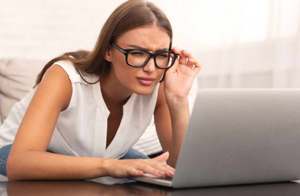 A young woman is squinting and trying to read her laptop while holding her glasses close to her face with her left hand
