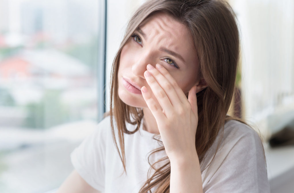 A young woman sitting by a window is gently touching her irritated eyes, experiencing the discomfort of eye strain.