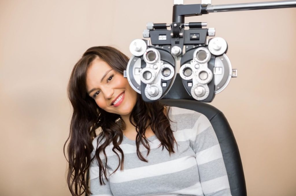 A woman smiling and sitting behind a phoropter at the optometrist's office