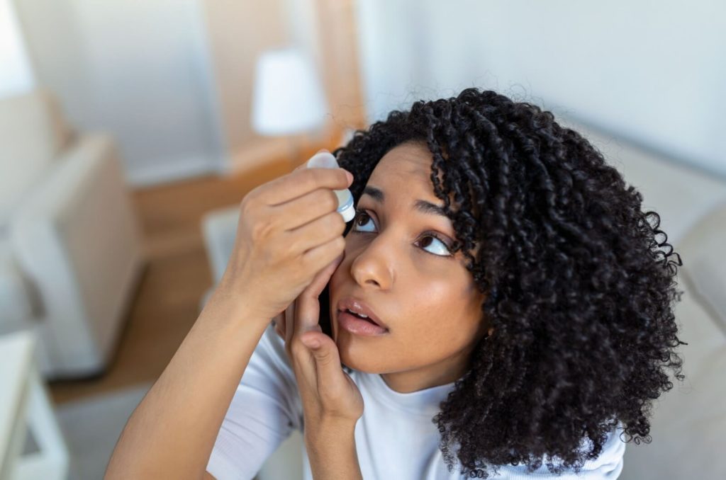 A woman looking up with her left hand under her right eye while putting eye drops with her right hand.