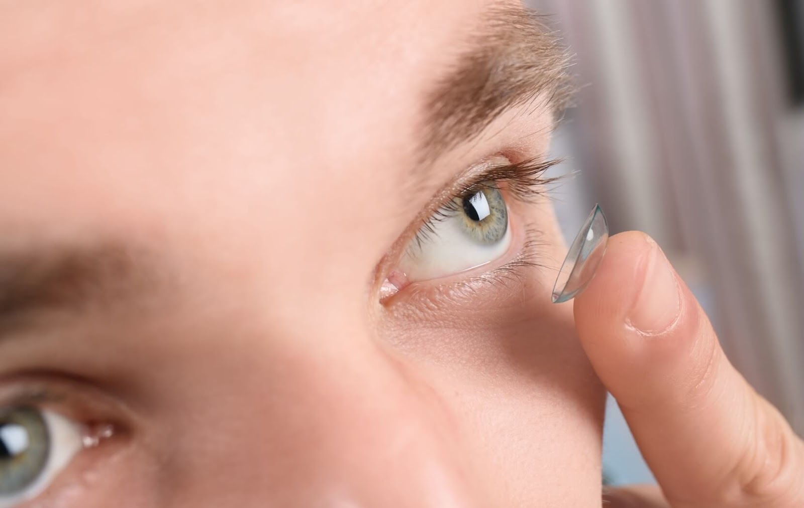 A person with light green eyes uses their finger to put a contact lens in their eye.