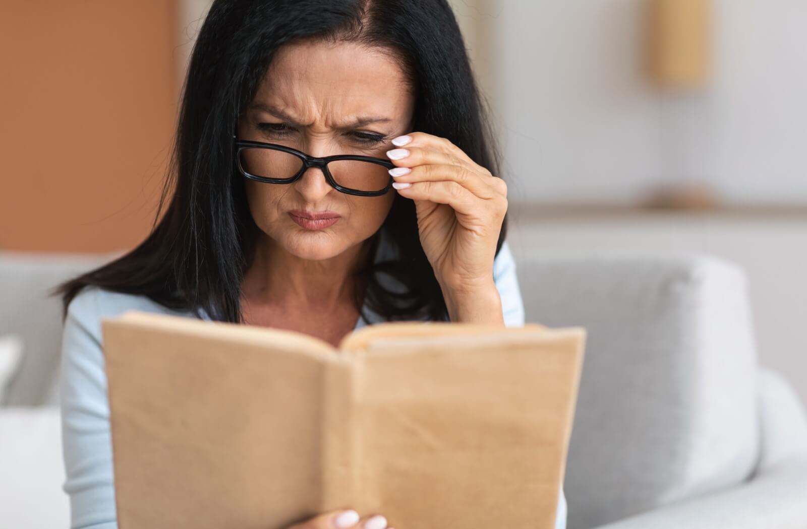 An adult squinting and adjusting their glasses while reading a book, showing signs of visual discomfort.