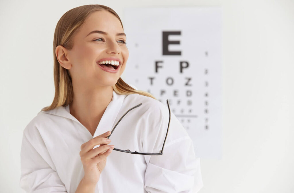 Smiling person in a white shirt holds eyeglasses in front of an eye chart during a vision test at an optometry clinic.