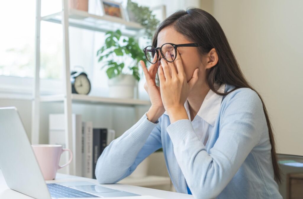 Woman wearing glasses rubbing tired eyes while sitting at a laptop at home.