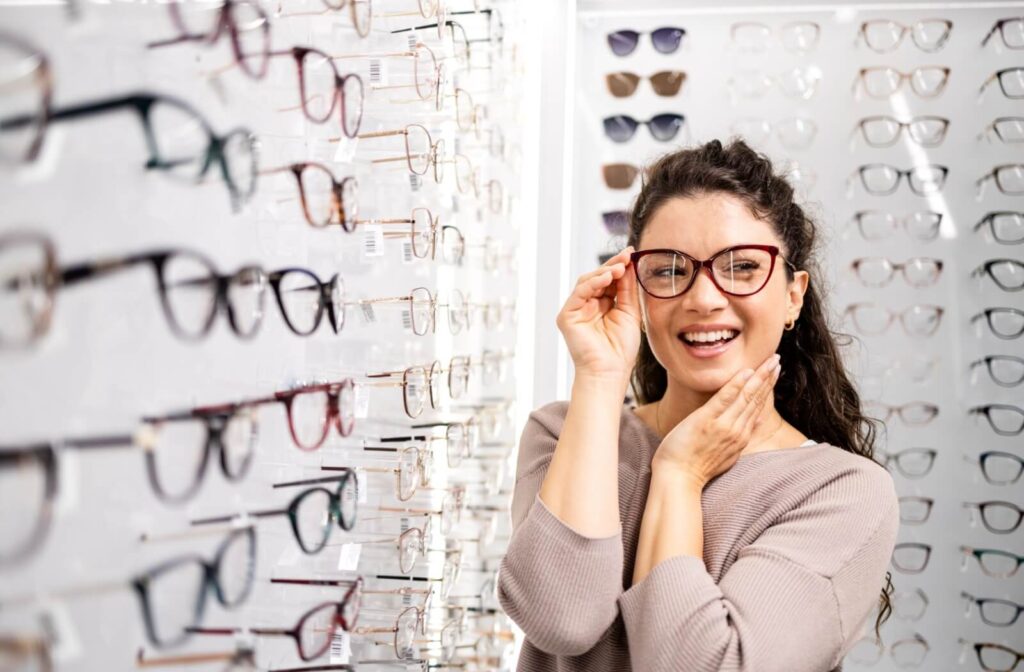 A smiling person with dark curly hair trying on dark red cat-eye glasses while standing in front of a large display of various eyeglass frames.
