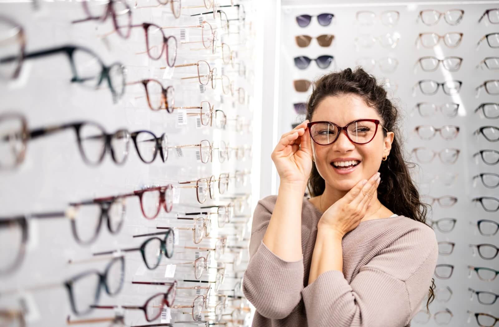 A smiling person with dark curly hair trying on dark red cat-eye glasses while standing in front of a large display of various eyeglass frames.
