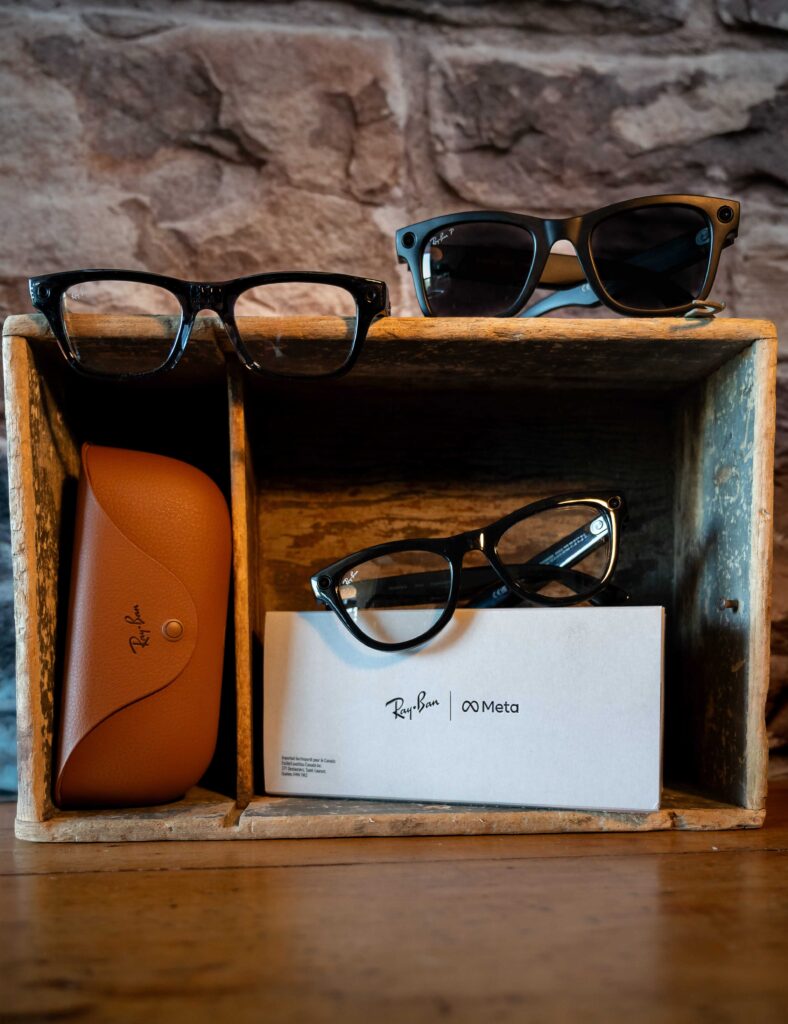 A retail display featuring multiple pairs of Ray-Ban Meta smart glasses with built-in cameras, shown alongside a brown leather charging case and branded white packaging box.
