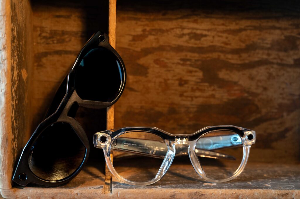 Two pairs of Ray-Ban Meta smart glasses displayed on a rustic wooden shelf, showcasing the contrast between the classic black sunglasses frame and the modern clear acetate frame options.