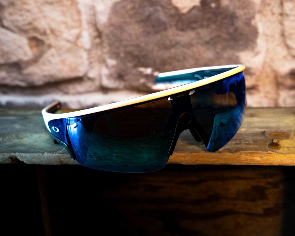 A close-up view of Oakley smart sunglasses featuring a white frame and a large, continuous blue shield lens, displayed on a rustic wooden shelf.