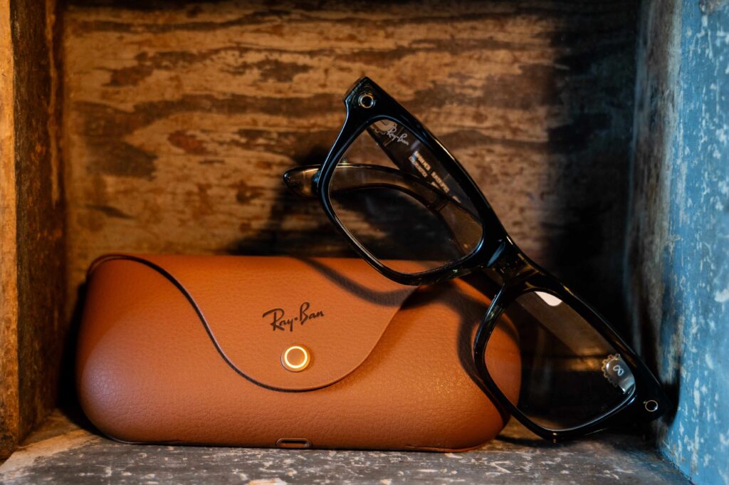 A pair of black Ray-Ban Meta smart prescription glasses leaning against their brown leather protective charging case on a rustic wooden shelf.